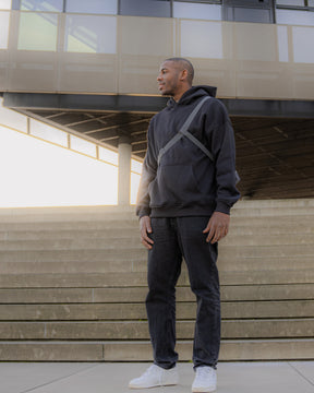 Mann trägt schwarze Umhängetasche 'Dylan' von Johnny Urban, schwarzen Hoodie, Jeans und weiße Sneaker vor modernen Treppen