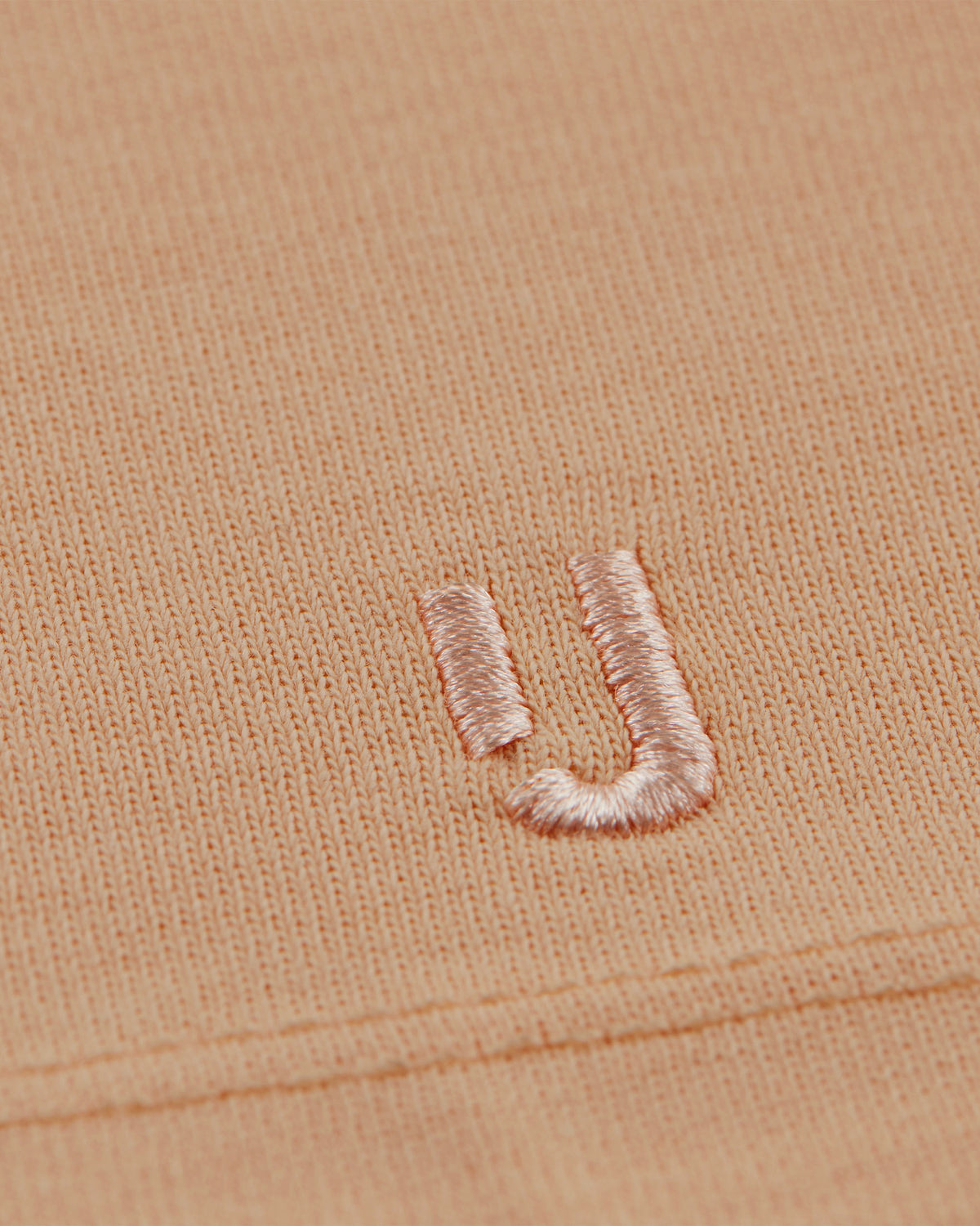 Nahaufnahme der rosa Stickerei des Buchstabens ‚JU‘ auf einem pfirsichfarbenen T-Shirt ‚Sammy‘ von Johnny Urban