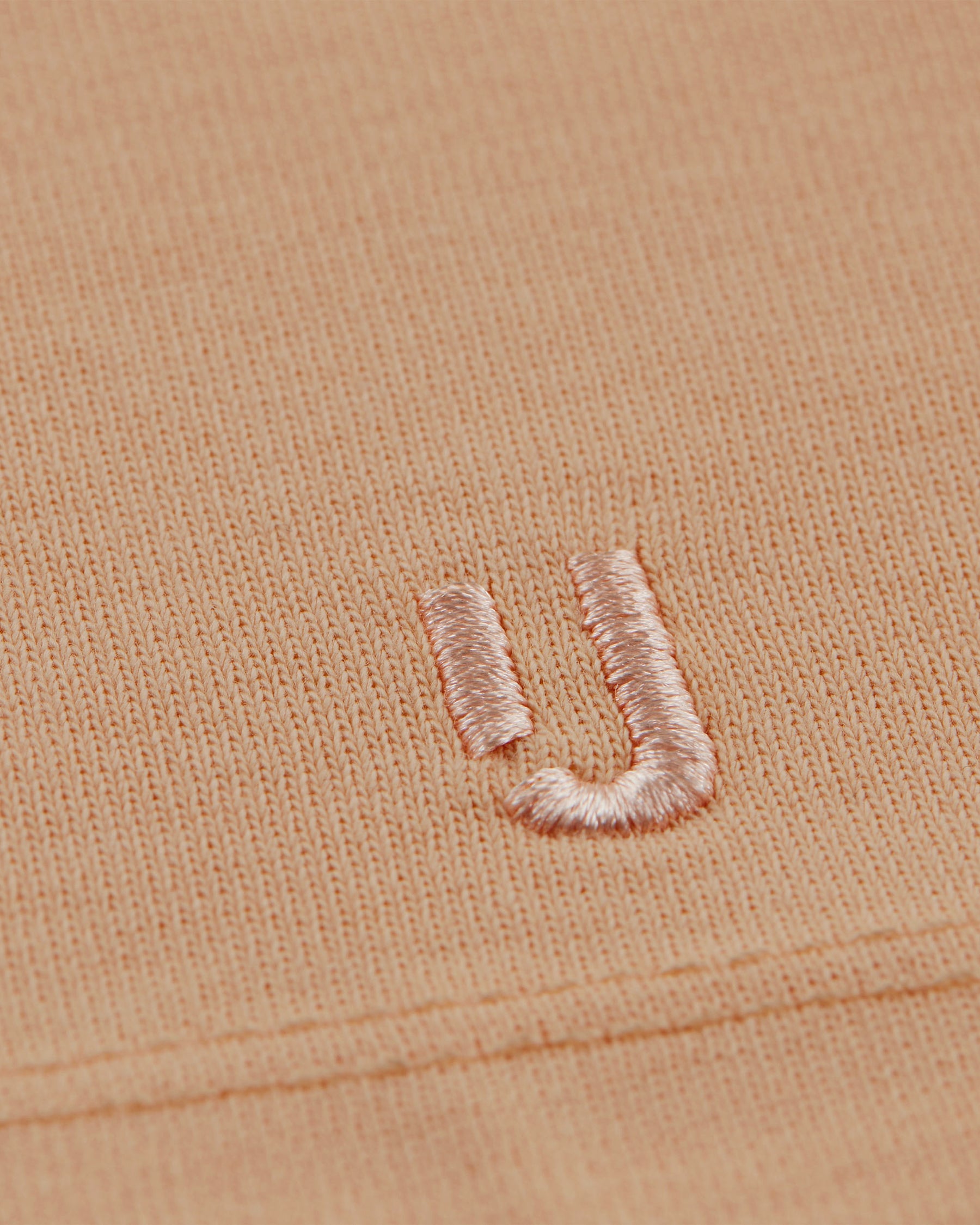 Nahaufnahme der rosa Stickerei des Buchstabens ‚JU‘ auf einem pfirsichfarbenen T-Shirt ‚Sammy‘ von Johnny Urban