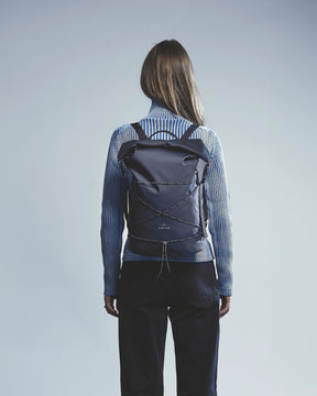 Rückansicht einer Frau mit langem braunem Haar, die einen schwarzen rolltop Rucksack 'Myles' von Johnny Urban und einen blauen Rollkragenpullover trägt