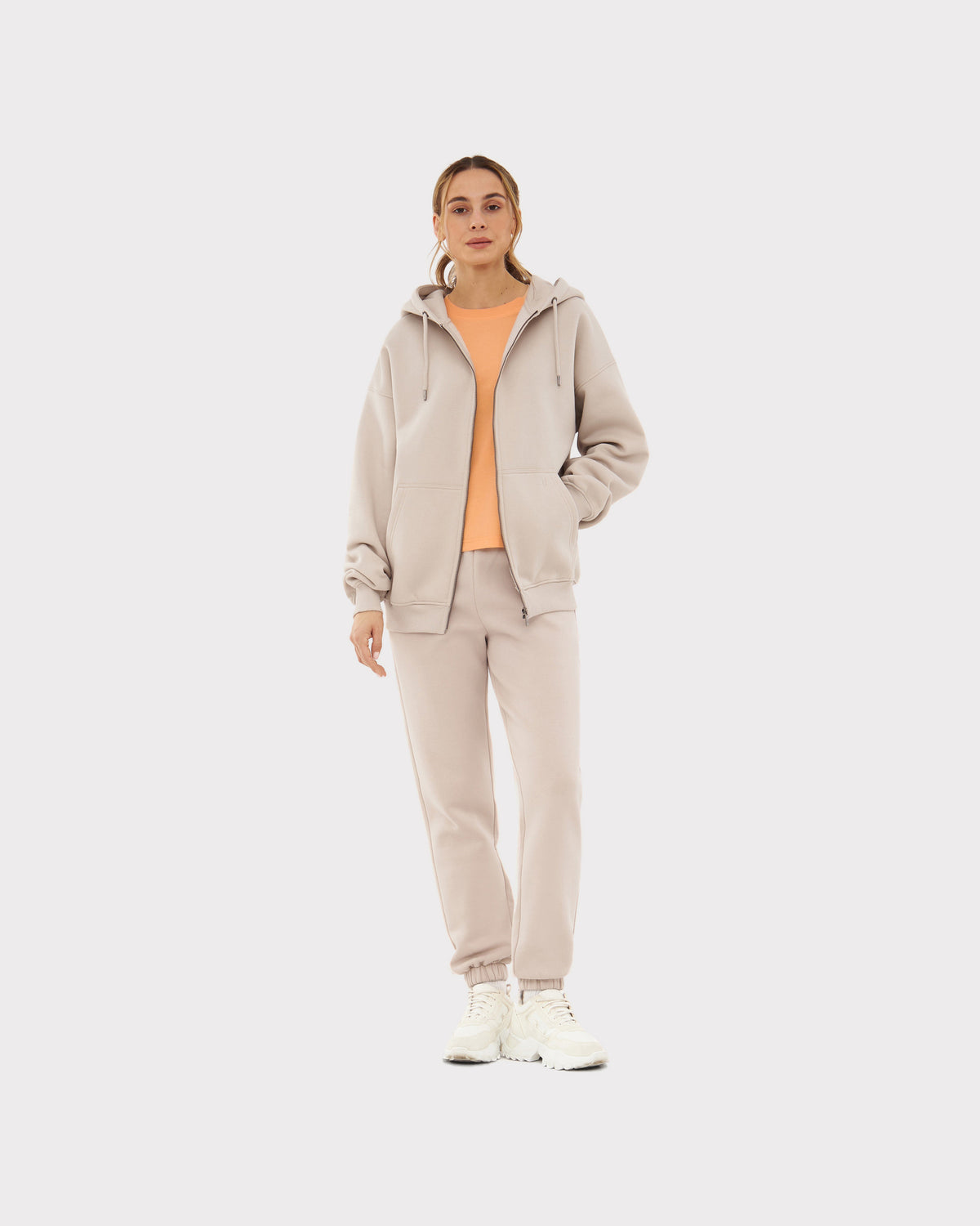 Frau trägt beige Oversized Zip Hoodie 'Caleb' mit Kapuze und bequeme Jogginghose, darüber orange T-Shirt