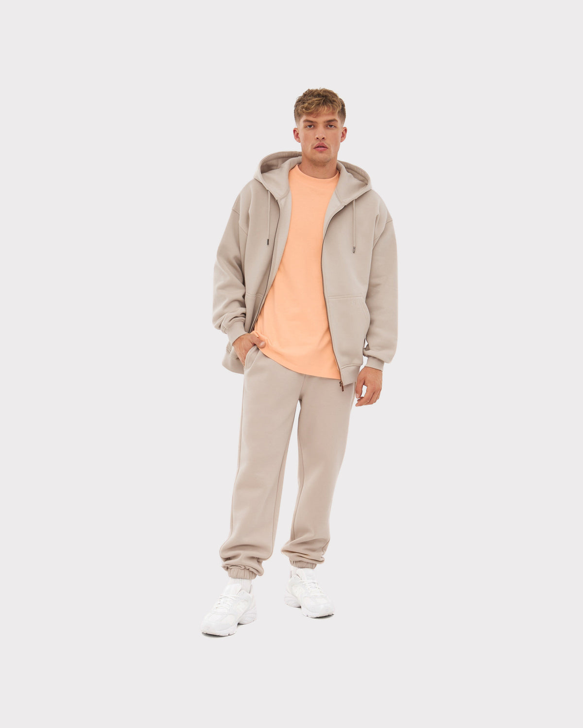 Mann trägt übergroße beige Zip-Hoodie 'Caleb' mit Kapuze, passende Jogginghose und weißer Sneaker