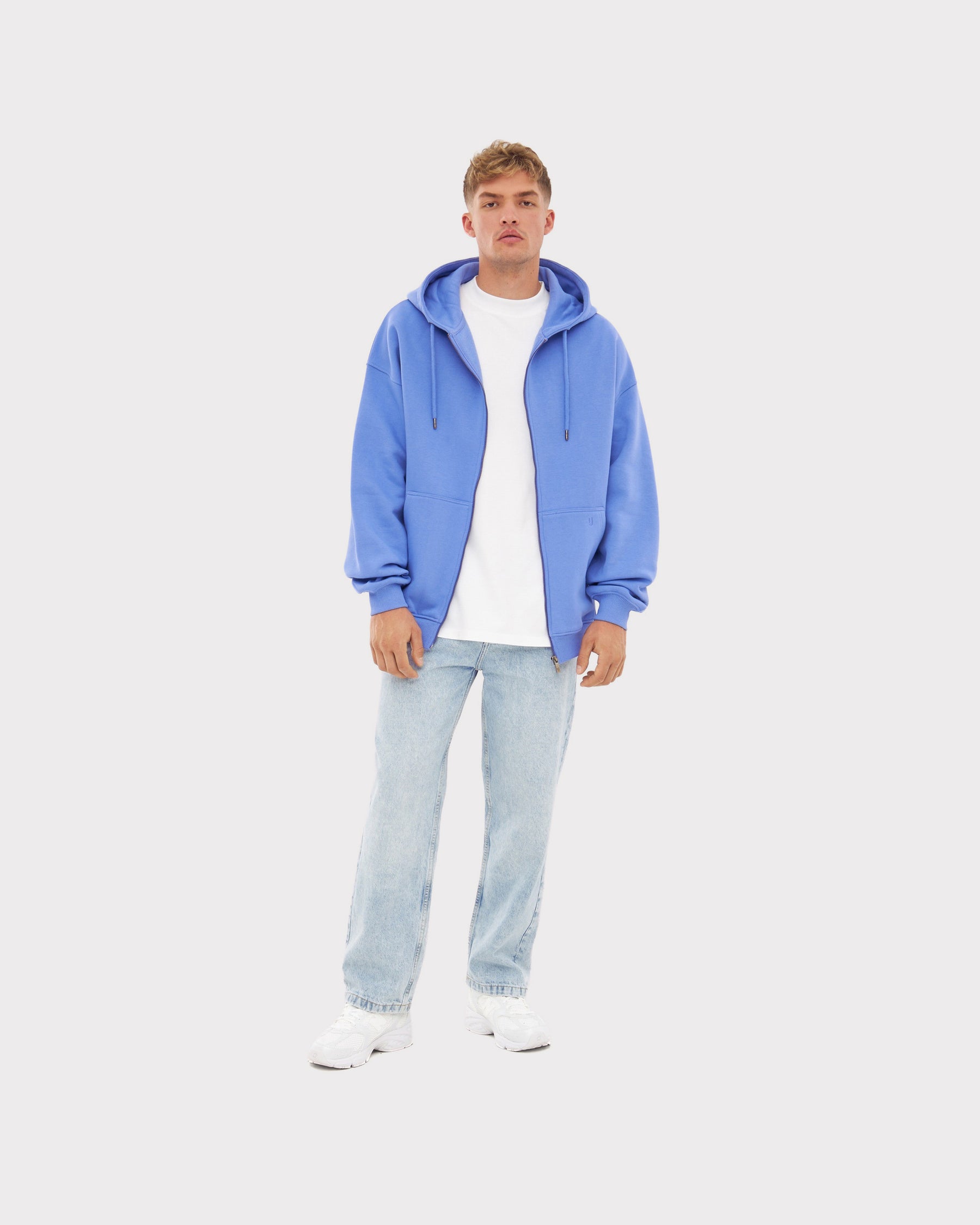 Mann trägt oversized Zip-Hoodie 'Caleb' in Blau-Lila, weißes T-Shirt, helle Jeans und weiße Schuhe