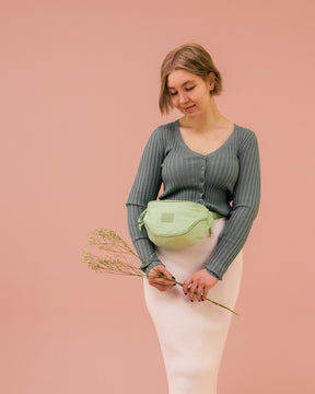 Frau mit grüner Bauchtasche ‚Ben‘ von Johnny Urban, grauer Strickjacke und weißem Rock vor rosa Hintergrund