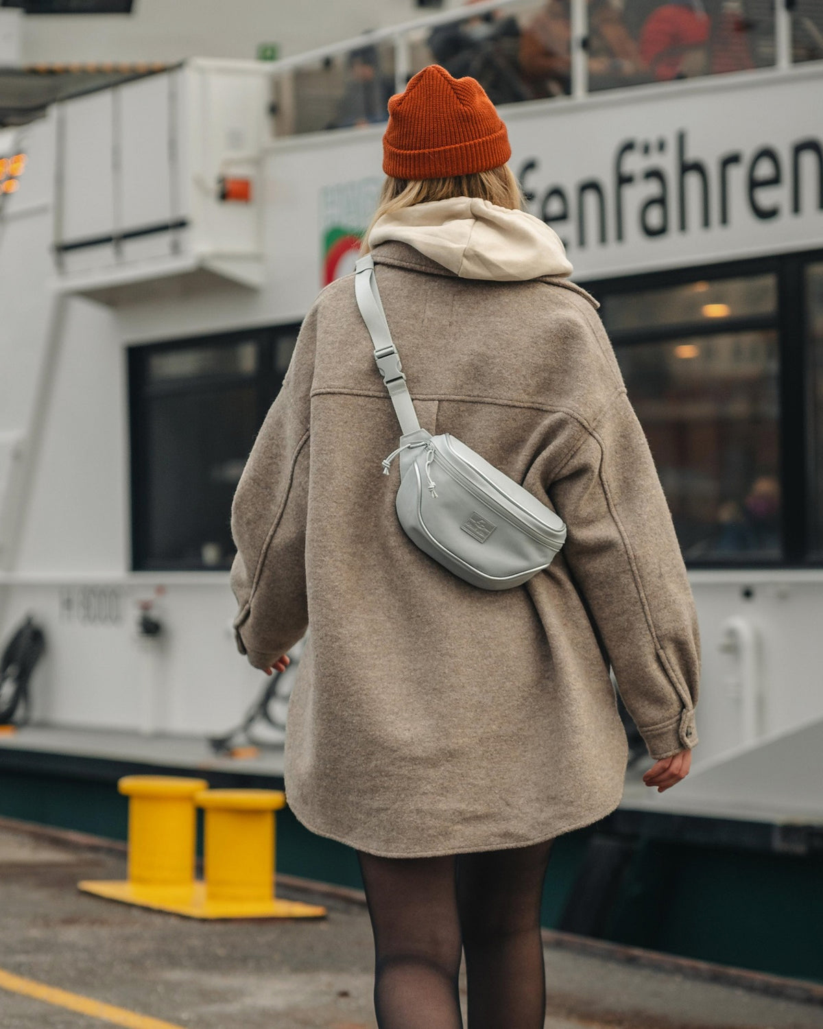 Dame trägt graue Johnny Urban Bauchtasche 'Ben' quer über dem Rücken, brauner Mantel, roter Strickmütze, im urbanen Hafenbereich