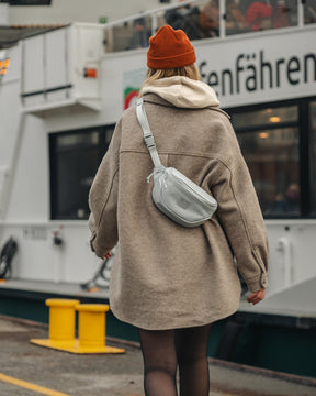 Dame trägt graue Johnny Urban Bauchtasche 'Ben' quer über dem Rücken, brauner Mantel, roter Strickmütze, im urbanen Hafenbereich