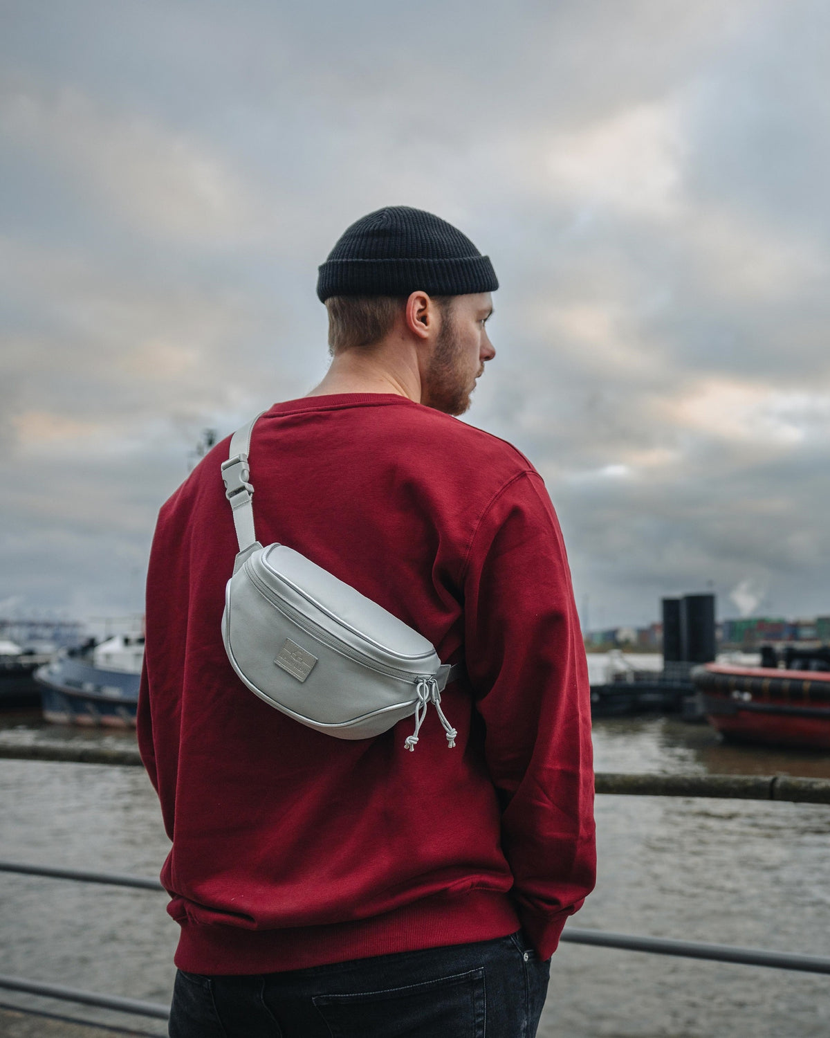 Mann trägt graue Bauchtasche 'Ben' von Johnny Urban über rotem Pullover am Hafen