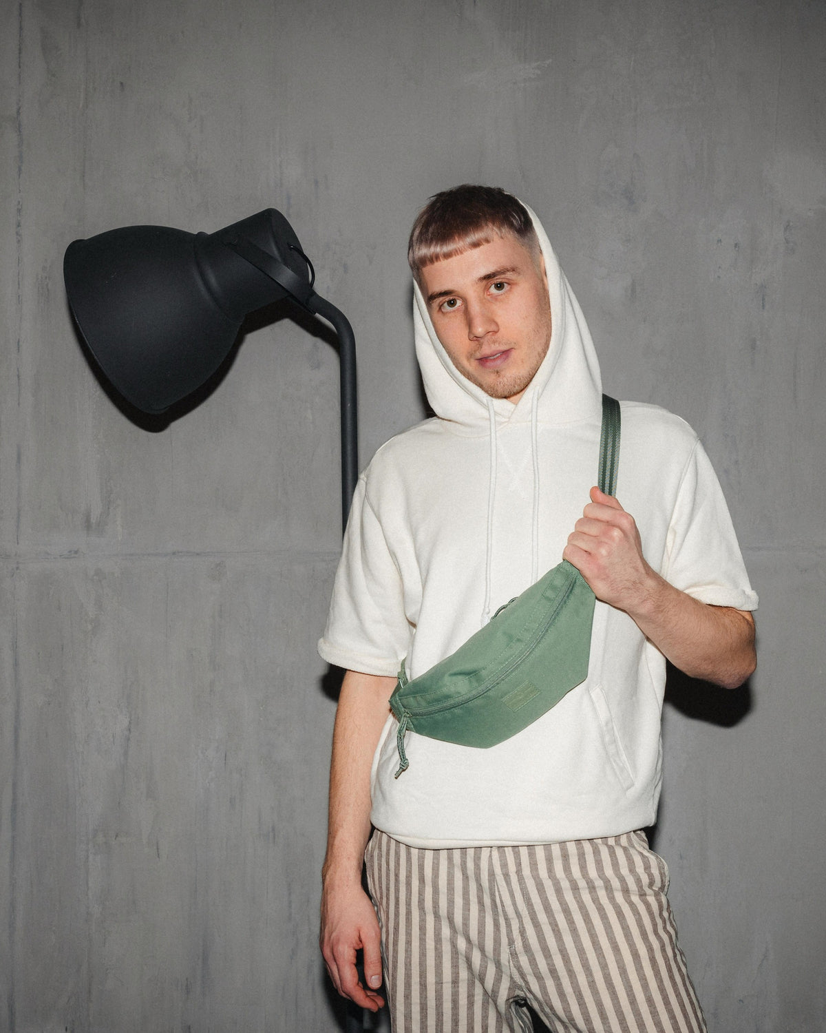 Mann trägt grüne Bauchtasche 'Erik Small' von Johnny Urban über der Schulter, weiße Kapuzenjacke, gestreifte Hose