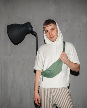 Mann trägt grüne Bauchtasche 'Erik Small' von Johnny Urban über der Schulter, weiße Kapuzenjacke, gestreifte Hose