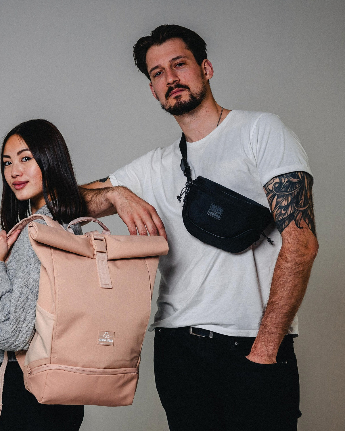 Mann trägt schwarze Bauchtasche 'Ben' von Johnny Urban quer über die Brust, Frau hält beigen Rucksack mit Rollverschluss