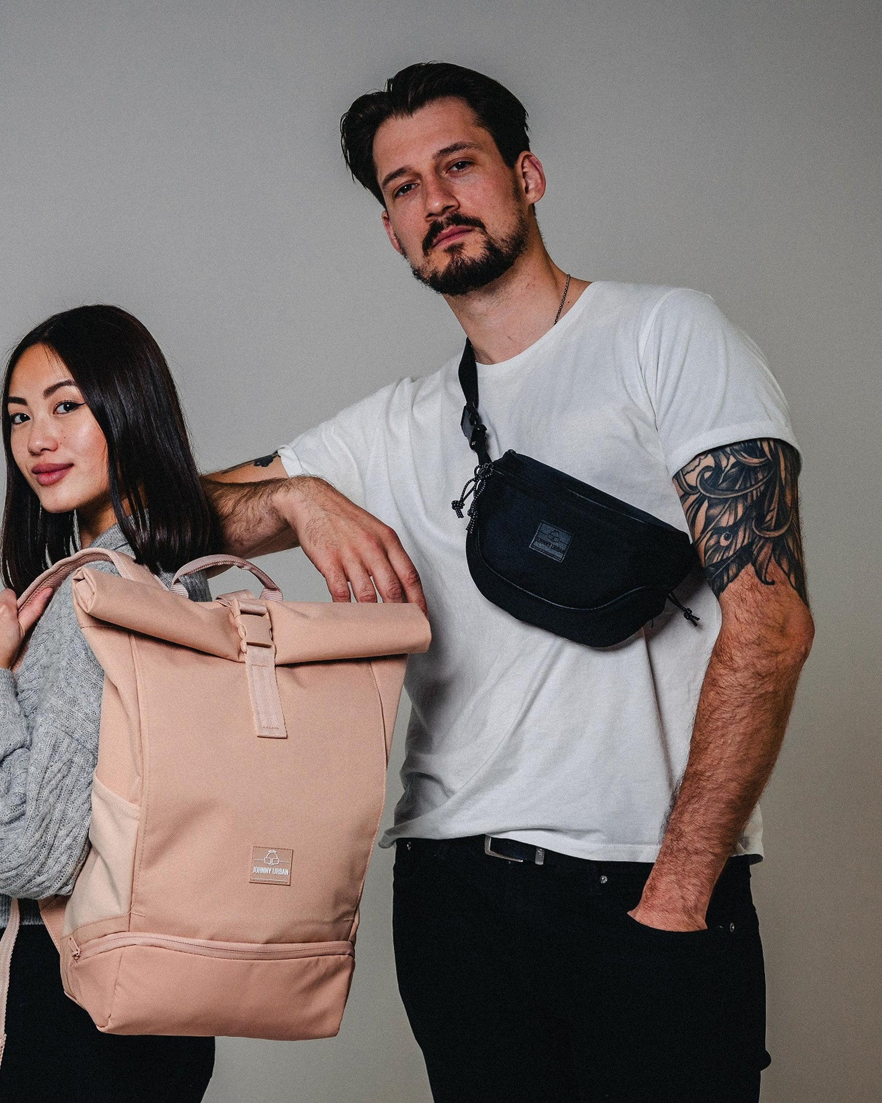 Mann trägt schwarze Bauchtasche 'Ben' von Johnny Urban quer über die Brust, Frau hält beigen Rucksack mit Rollverschluss
