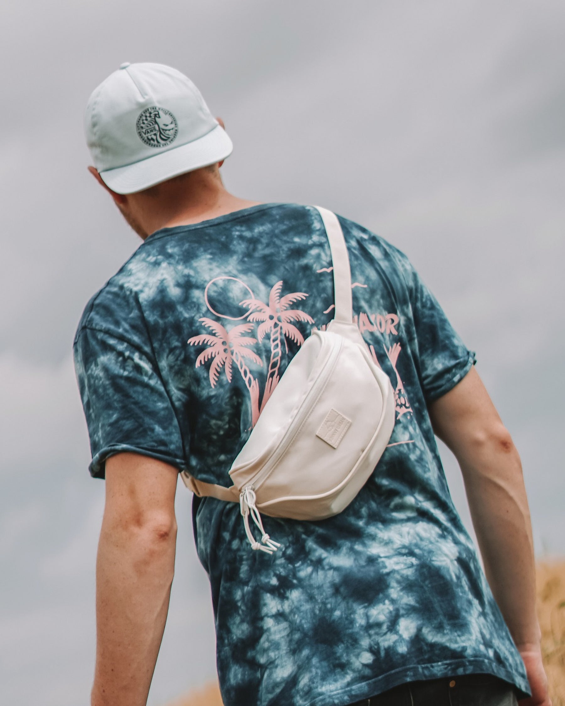 Mann von hinten trägt weiße Bauchtasche 'Ben' von Johnny Urban quer über den Rücken, dazu blau-schwarzes Batik-T-Shirt mit pinken Palmen