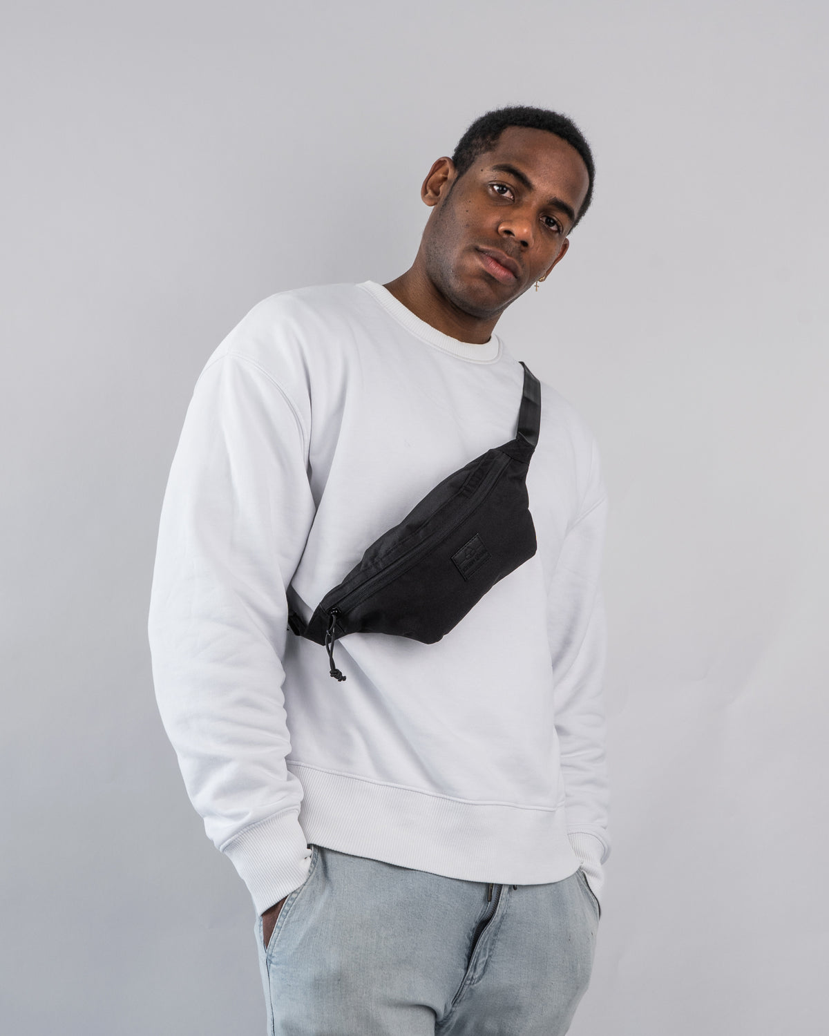 Mann trägt schwarze Johnny Urban Bauchtasche 'Erik Small' quer über der Brust mit weißem Pullover und Jeans