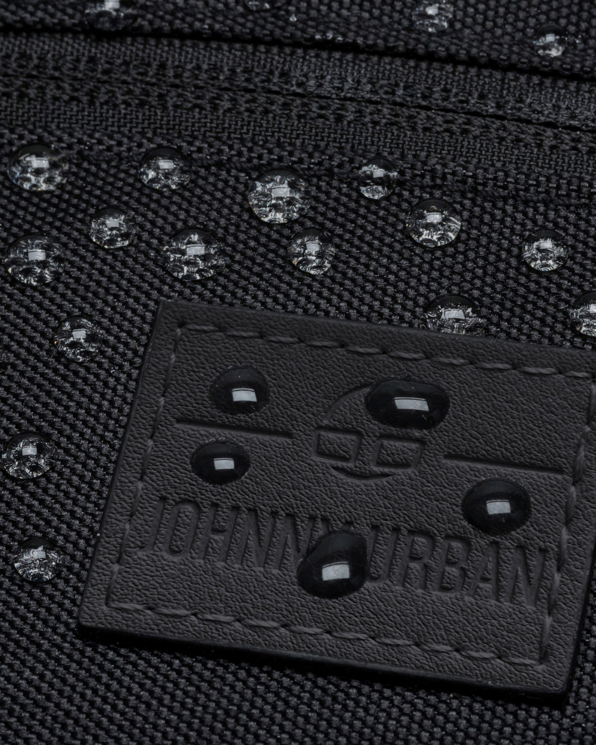 Nahaufnahme der wasserabweisenden schwarzen Bauchtasche 'Ben' mit geprägtem Johnny Urban Logo und Wassertropfen
