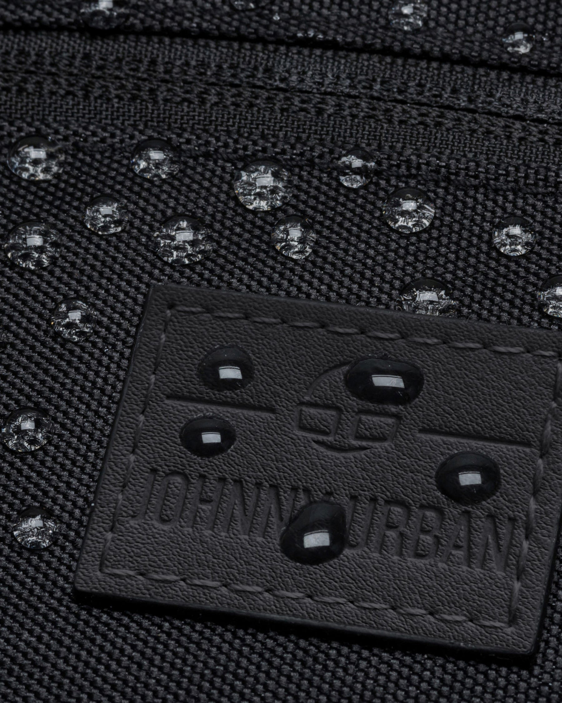 Nahaufnahme der wasserabweisenden schwarzen Bauchtasche 'Ben' mit geprägtem Johnny Urban Logo und Wassertropfen