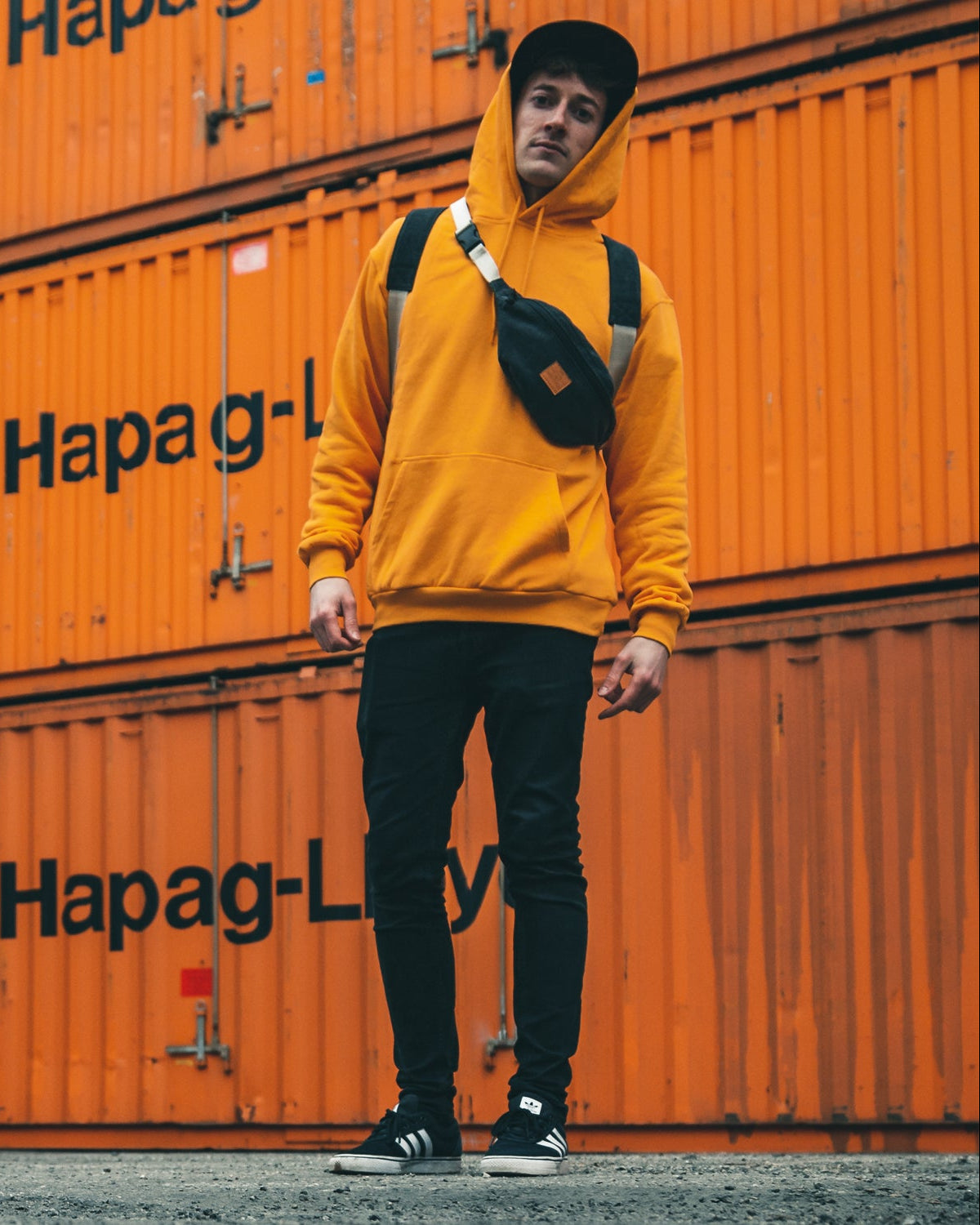 Mann trägt orangenen Hoodie, schwarze Hose, schwarze Cap und anthrazitfarbene Bauchtasche 'Toni' von Johnny Urban vor orangefarbenen Containern