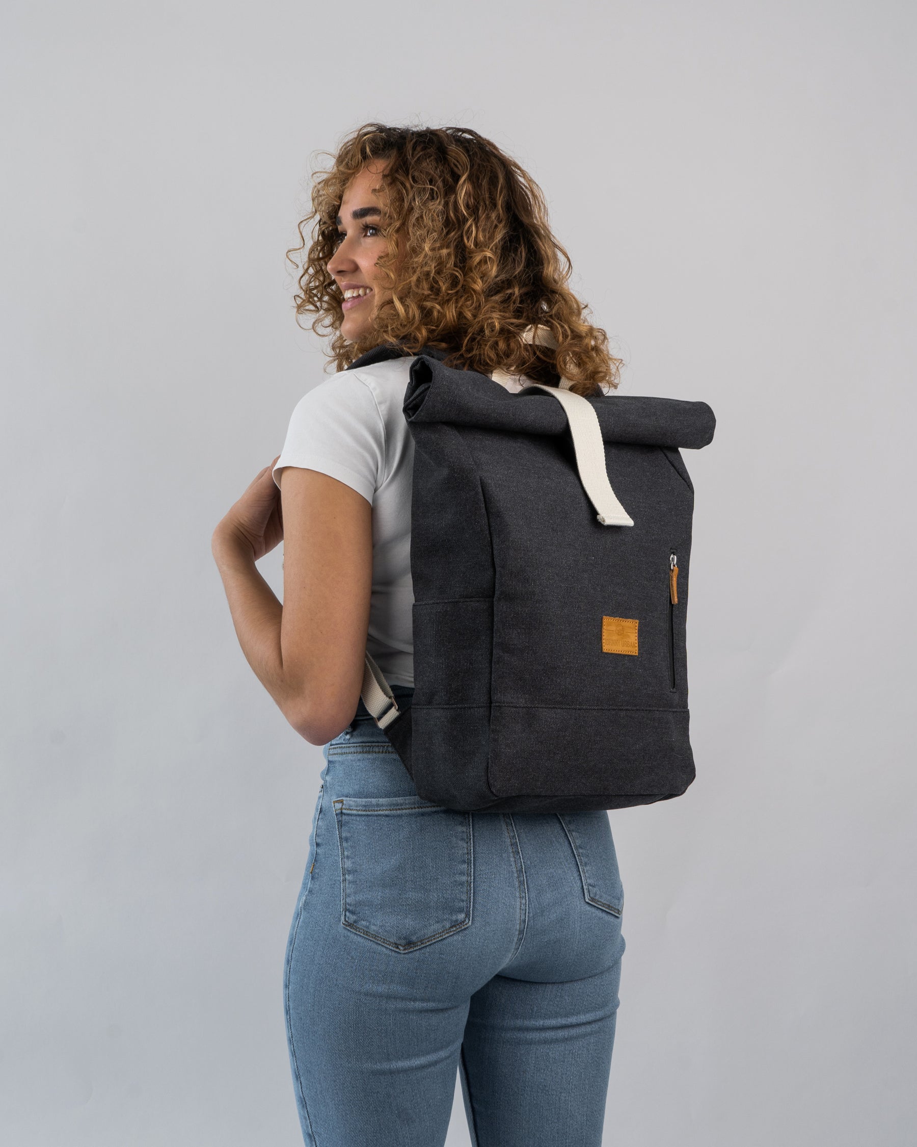 Frau mit lockigen Haaren trägt schwarzen Rolltop Rucksack 'Adam Medium' von Johnny Urban, helles T-Shirt, blaue Jeans