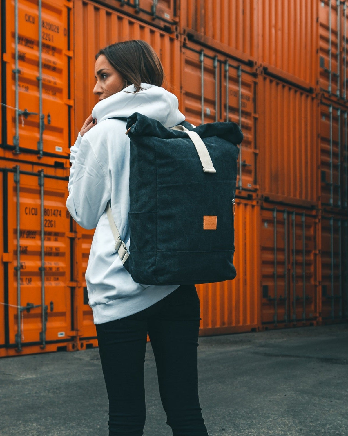 Frau trägt anthrazitfarbenen Rolltop Rucksack 'Adam Large' aus Canvas vor orangefarbenen Containern