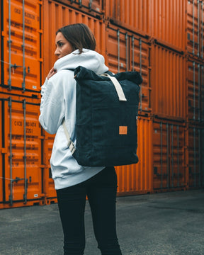 Frau trägt anthrazitfarbenen Rolltop Rucksack 'Adam Large' aus Canvas vor orangefarbenen Containern