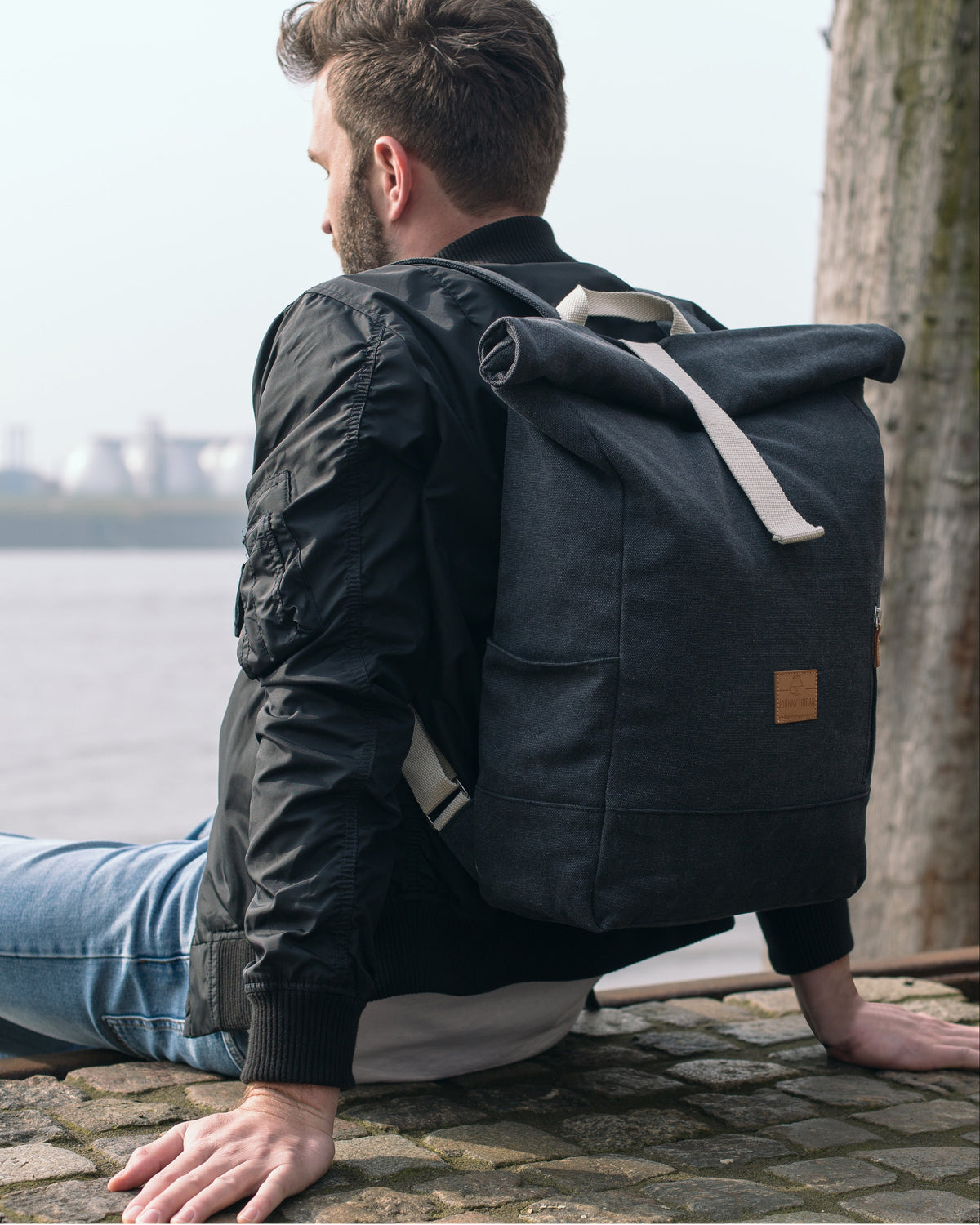Mann sitzt an Ufer mit anthrazitfarbenem Rolltop Rucksack „Adam Large“ aus Canvas