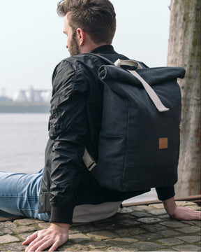 Mann sitzt an Ufer mit anthrazitfarbenem Rolltop Rucksack „Adam Large“ aus Canvas