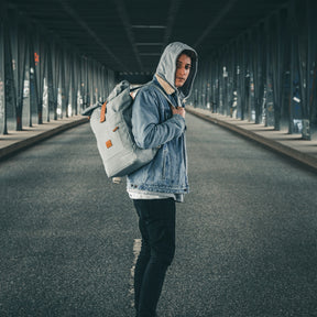 Junger Mann trägt grauen Rolltop Rucksack 'Adam Large' und Jeansjacke mit Kapuzenpullover unter einer Brücke
