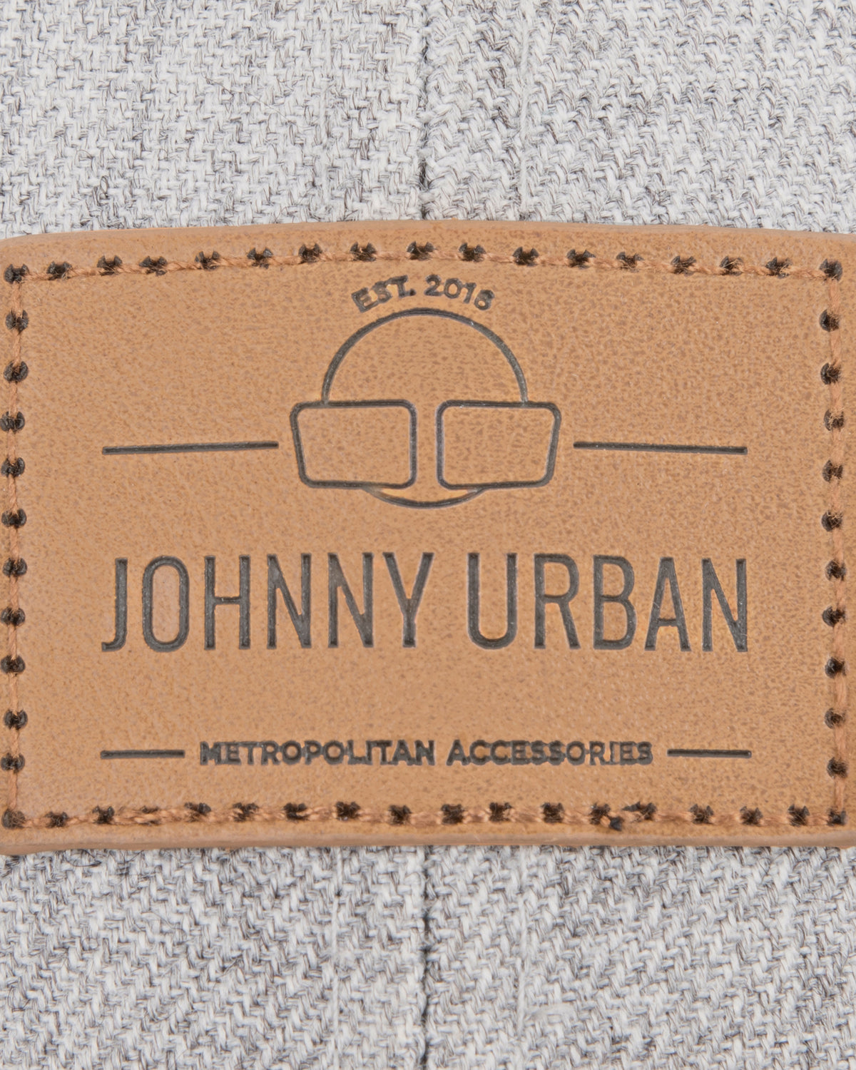 Nahaufnahme eines braunen Lederetiketts mit dem Schriftzug 'JOHNNY URBAN METROPOLITAN ACCESSORIES' auf graubraunem Stoff