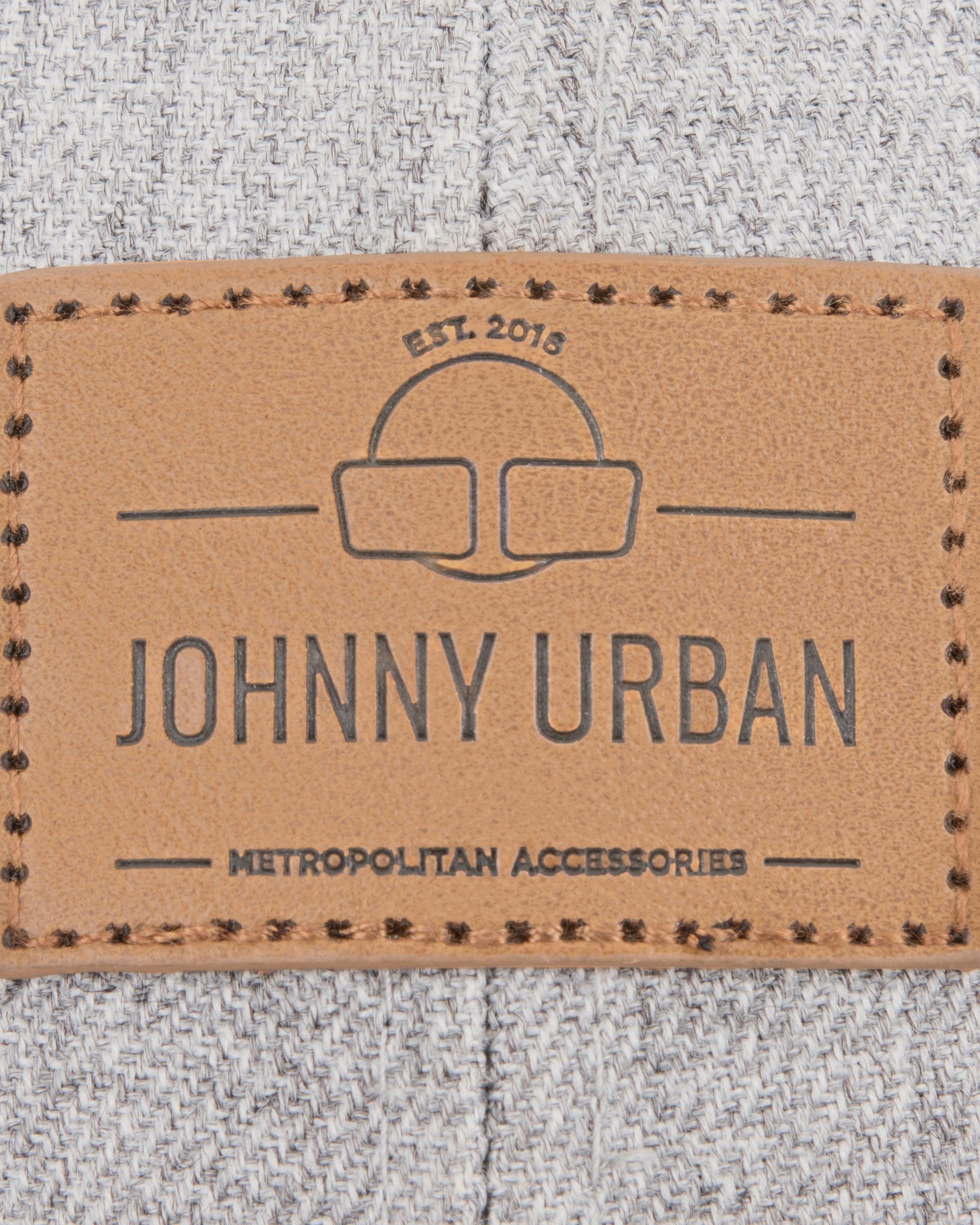 Nahaufnahme eines braunen Lederetiketts mit dem Schriftzug 'JOHNNY URBAN METROPOLITAN ACCESSORIES' auf graubraunem Stoff