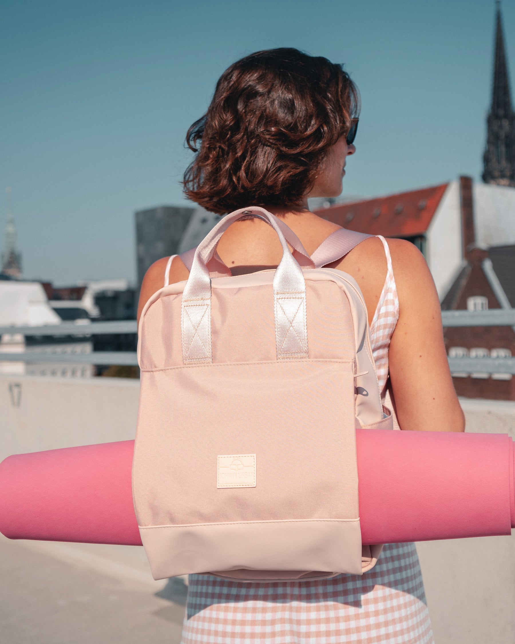 Rucksack 'Jona Large' in Rosa mit integriertem Tragegurt hält eine Yogamatte, getragen von Frau in ärmellosem Kleid