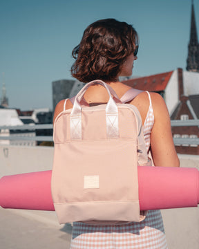 Rucksack 'Jona Large' in Rosa mit integriertem Tragegurt hält eine Yogamatte, getragen von Frau in ärmellosem Kleid