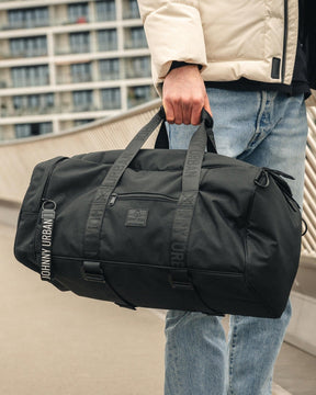Schwarze Johnny Urban Duffle Bag 'Colin' aus robustem Material, getragen von Mann in beiger Jacke und Jeans