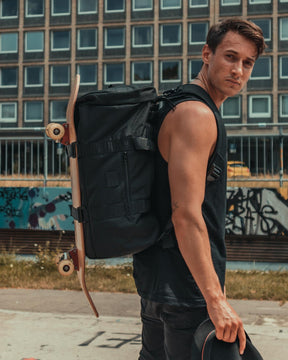 Mann mit schwarzem Duffle Bag 'Colin' und Skateboard an Rücken, hält schwarzen Hut, urbaner Hintergrund