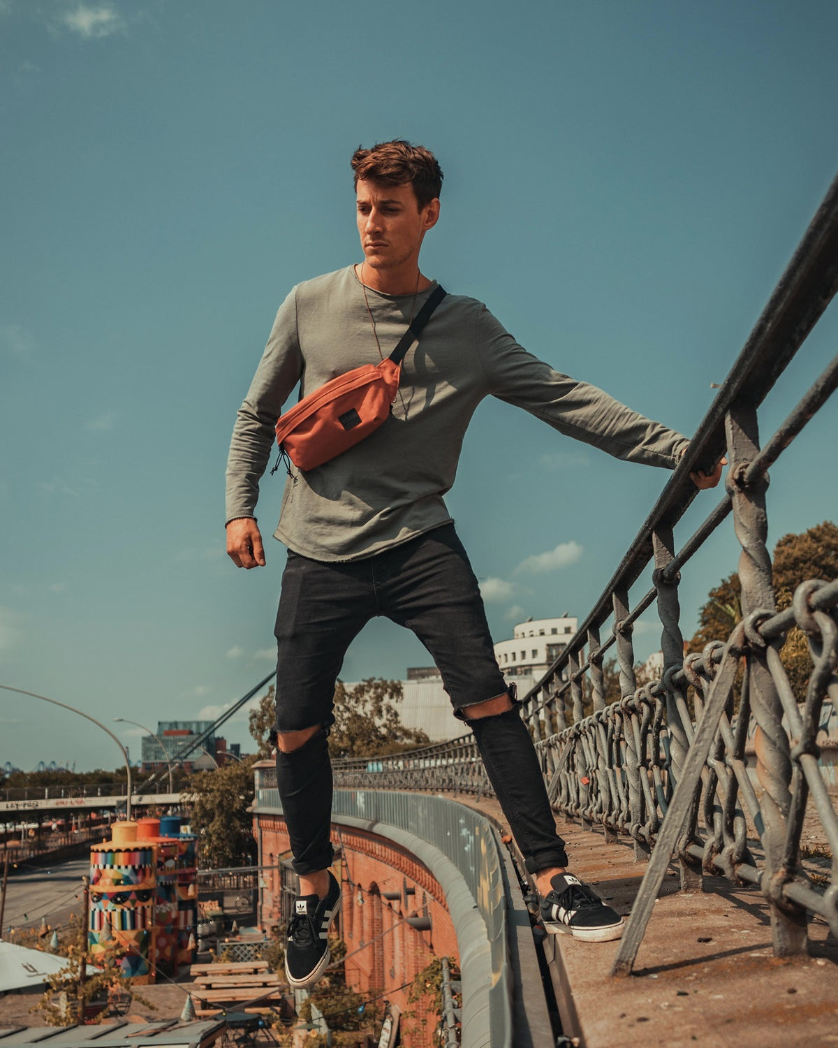 Mann trägt orangefarbene Bauchtasche 'Tom', graues Langarmshirt, schwarze zerrissene Jeans und schwarze Sneaker auf Brückengeländer