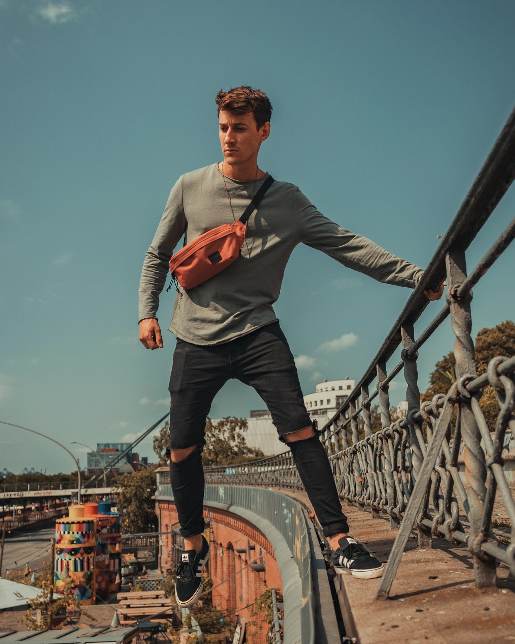 Mann trägt orangefarbene Bauchtasche 'Tom', graues Langarmshirt, schwarze zerrissene Jeans und schwarze Sneaker auf Brückengeländer