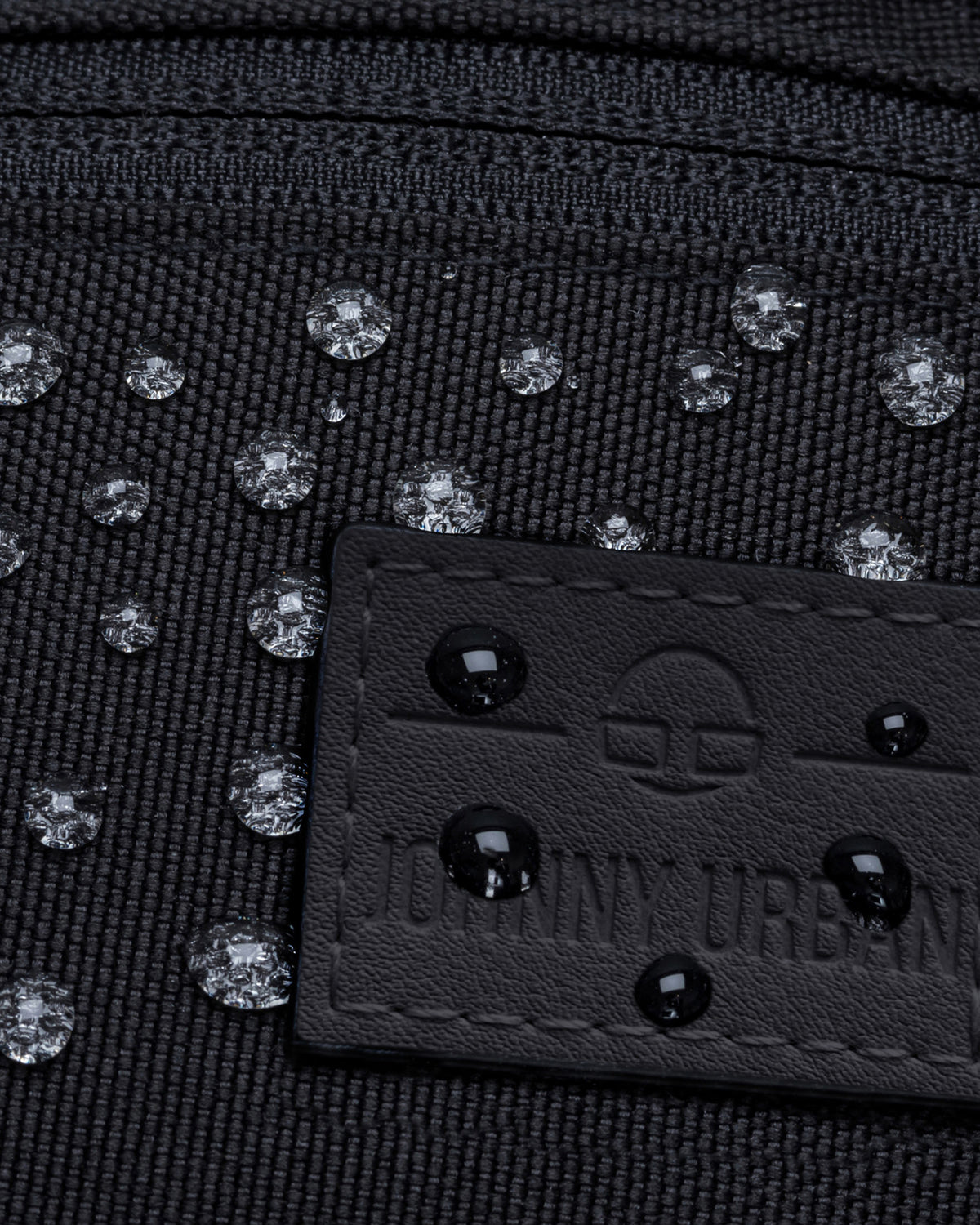 Nahe Aufnahme einer wasserabweisenden schwarzen Bauchtasche 'Erik Small' mit Wassertropfen auf dem Stoff und dem Johnny Urban Label