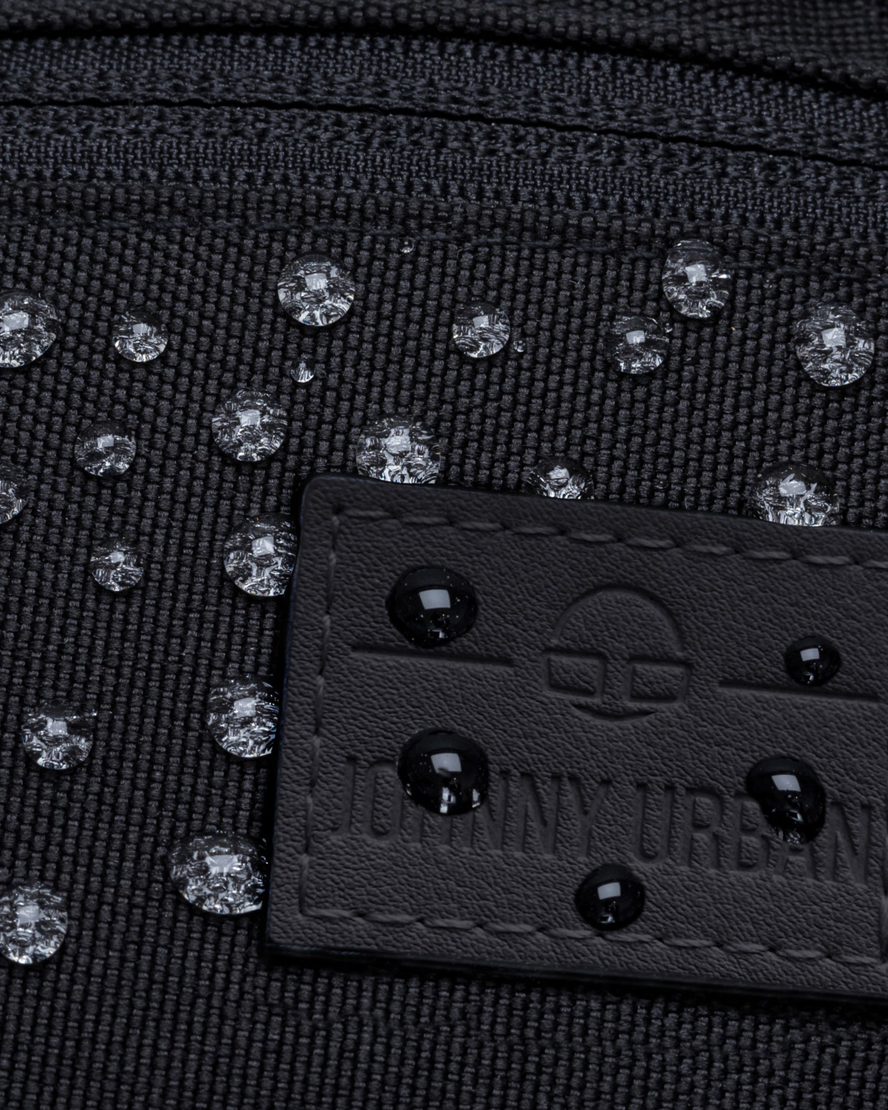 Nahe Aufnahme einer wasserabweisenden schwarzen Bauchtasche 'Erik Small' mit Wassertropfen auf dem Stoff und dem Johnny Urban Label