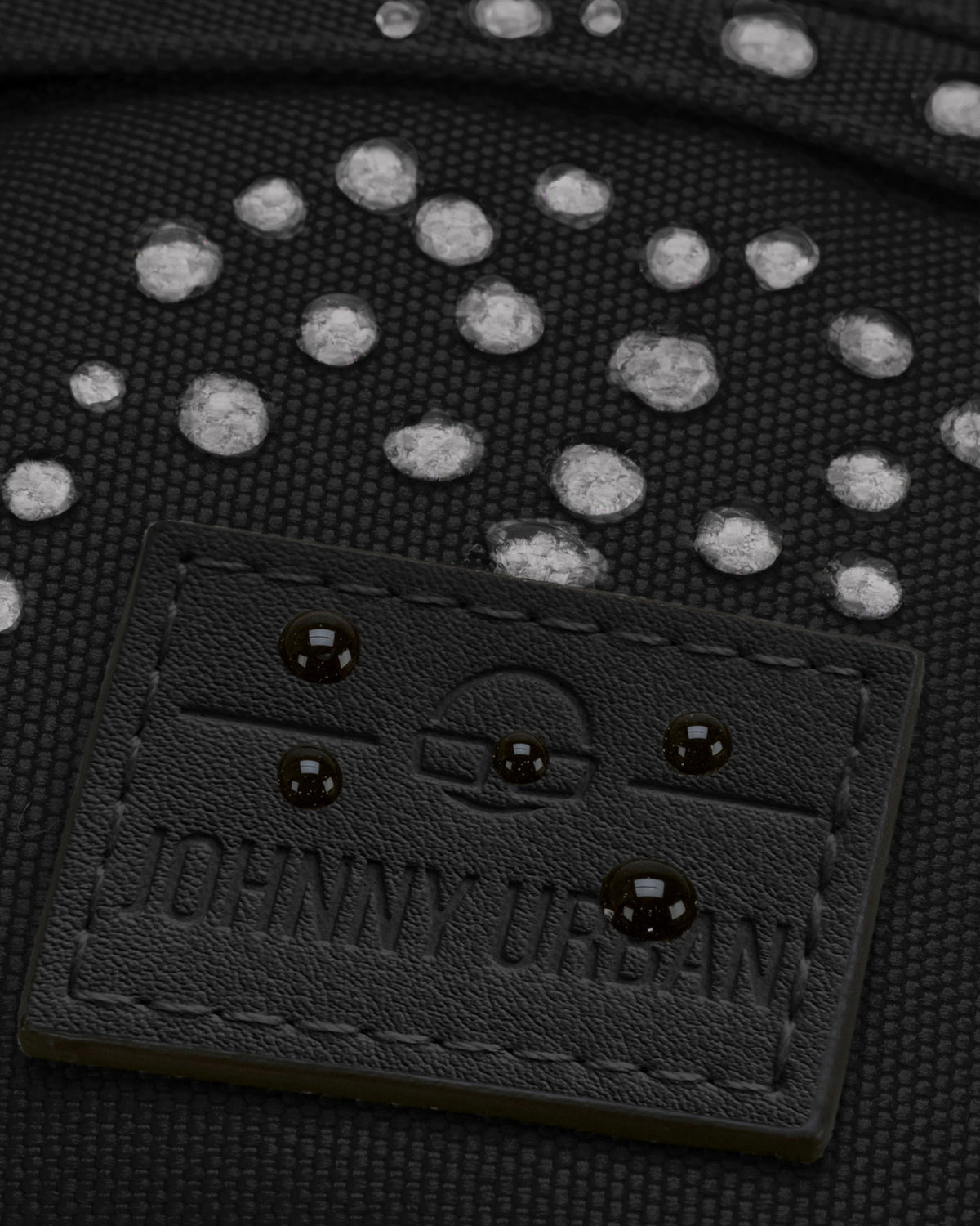 Nahaufnahme der wasserabweisenden schwarzen Bauchtasche 'Tom' von Johnny Urban mit sichtbaren Wassertropfen auf dem Material und geprägtem Logo