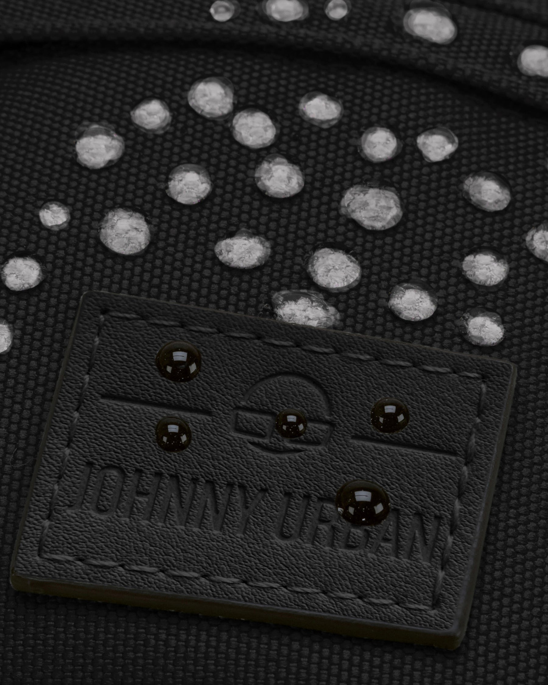 Nahaufnahme der wasserabweisenden schwarzen Bauchtasche 'Tom' von Johnny Urban mit sichtbaren Wassertropfen auf dem Material und geprägtem Logo