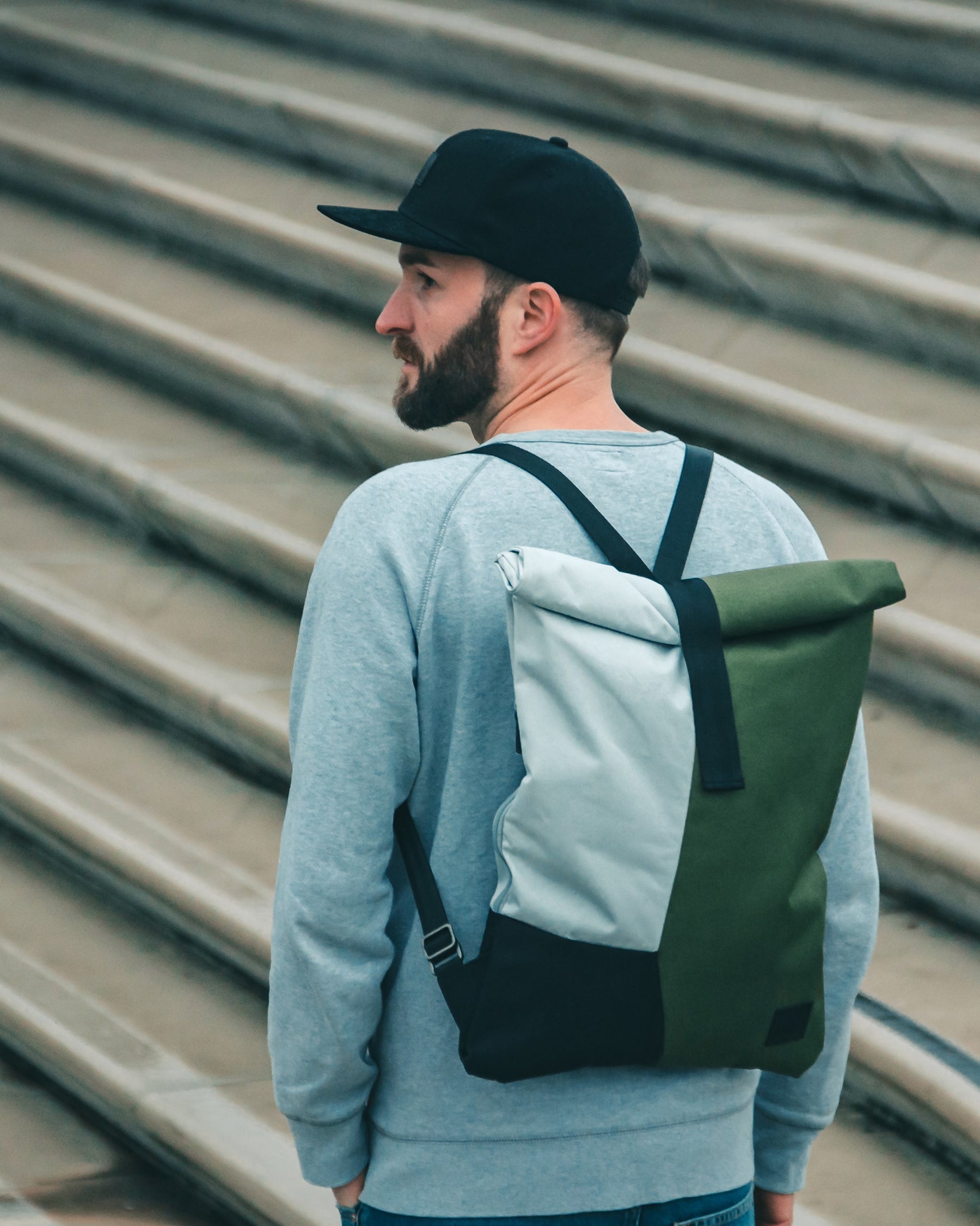 Mann mit schwarzer Cap 'Dean', grauem Pullover und zweifarbigen Rucksack vor einer Treppe
