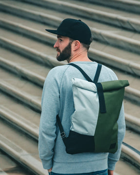 Mann mit schwarzer Cap 'Dean', grauem Pullover und zweifarbigen Rucksack vor einer Treppe