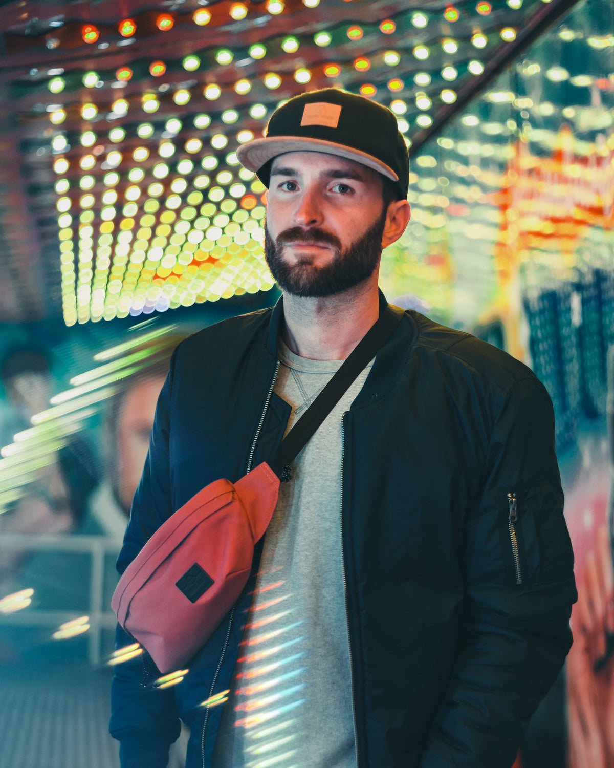Mann mit schwarzer 'Dean'-Cap, roter Gürteltasche und schwarzer Jacke vor leuchtender urbaner Kulisse