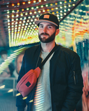 Mann mit schwarzer 'Dean'-Cap, roter Gürteltasche und schwarzer Jacke vor leuchtender urbaner Kulisse