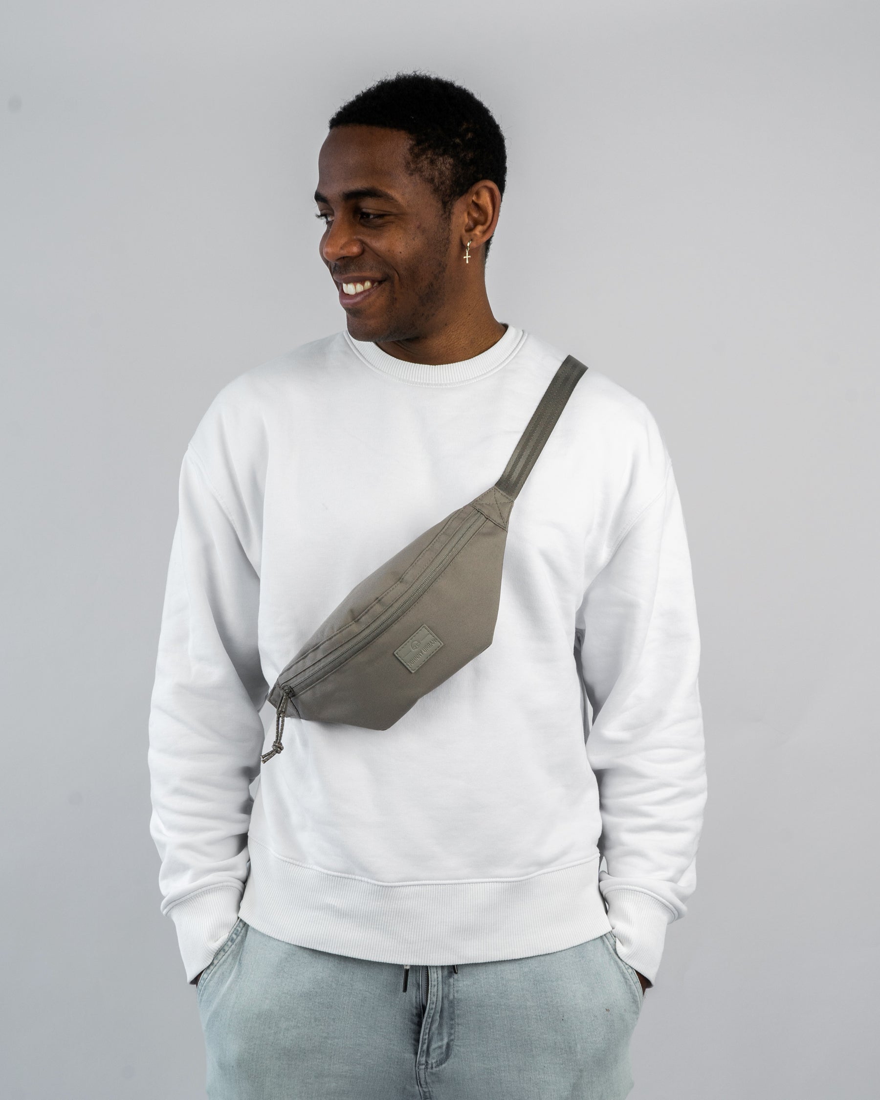 Mann trägt die Johnny Urban Bauchtasche 'Erik Small' in Grau über der Brust, kombiniert mit weißem Sweatshirt und hellblauen Jeans