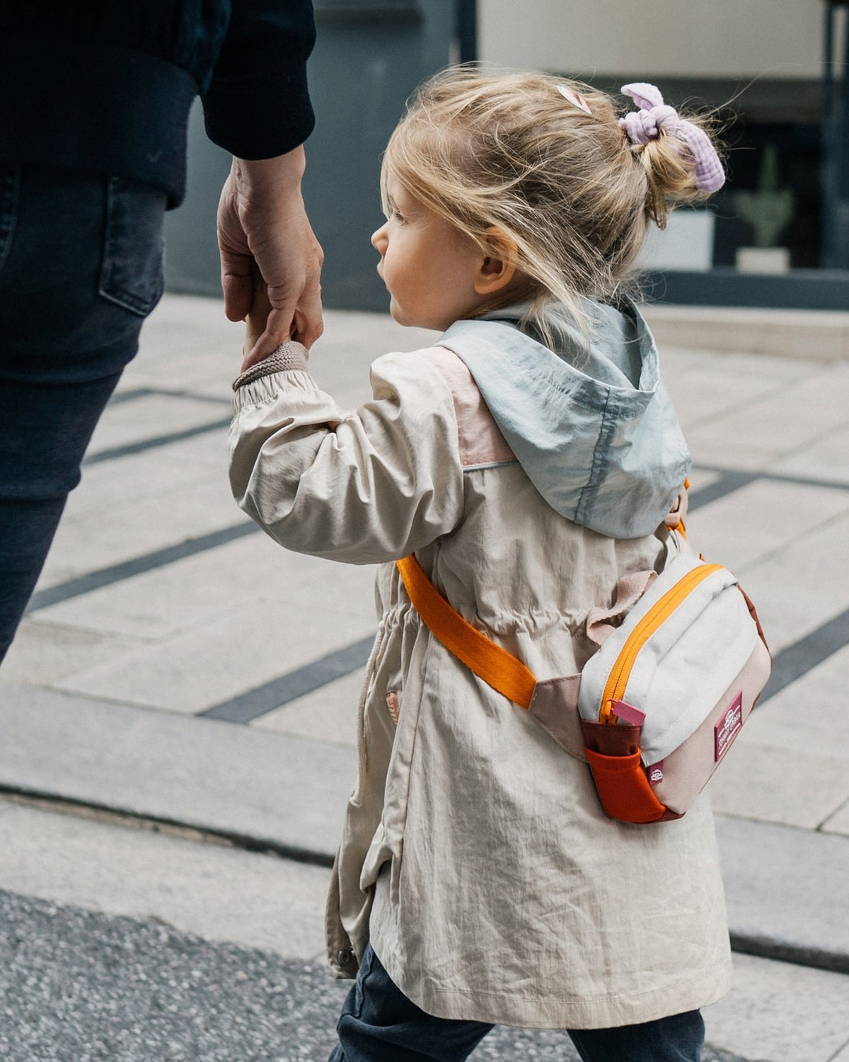 Kindermädchen mit hochgestecktem Haar hält die Hand eines Erwachsenen und trägt die Kinder Bauchtasche 'Felix' in Beige mit orangefarbenen Akzenten