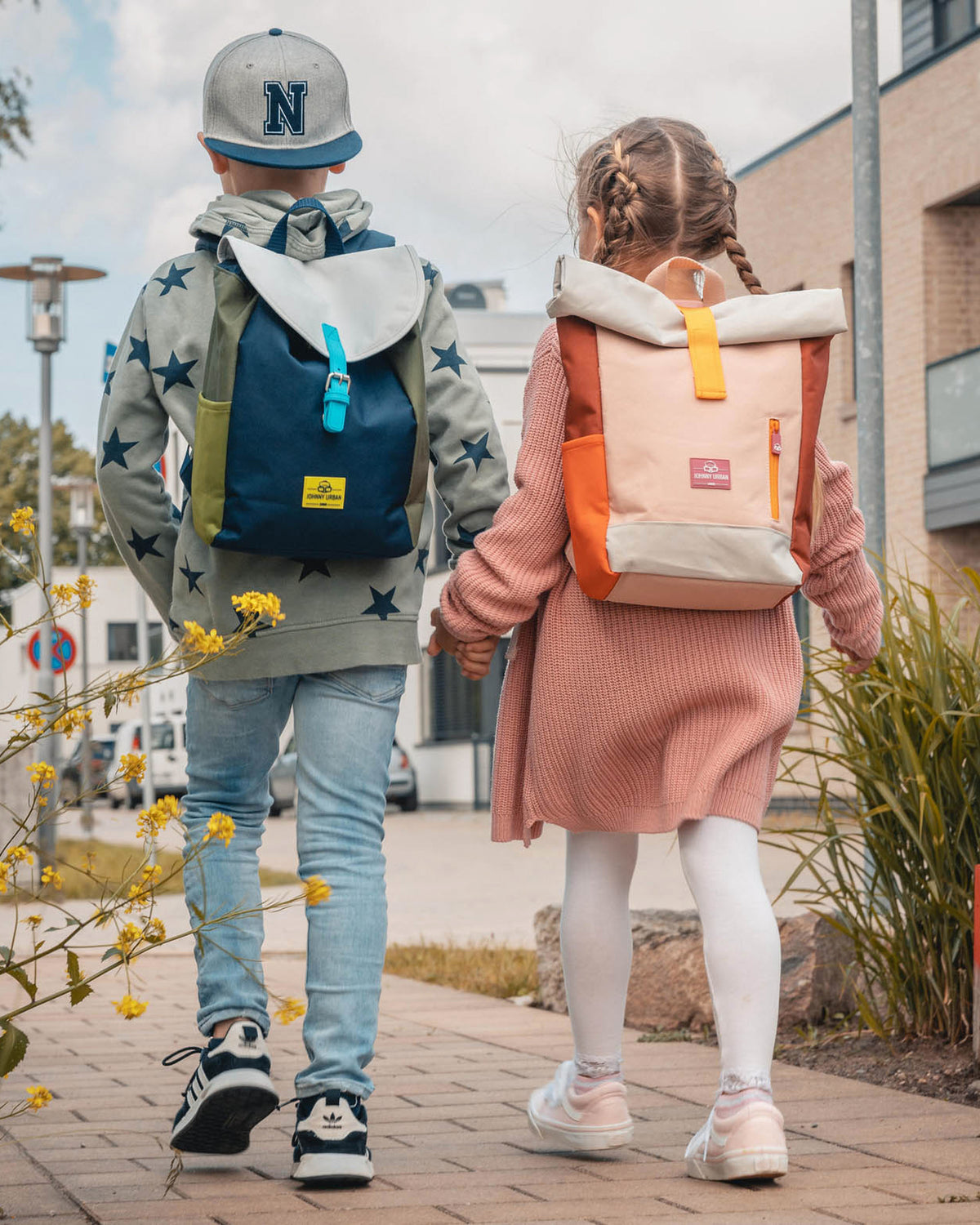 Junge mit blau-grünem und Mädchen mit rosa-orangefarbenem Kinderrucksack 'Liam' gehen Hand in Hand auf einem Gehweg
