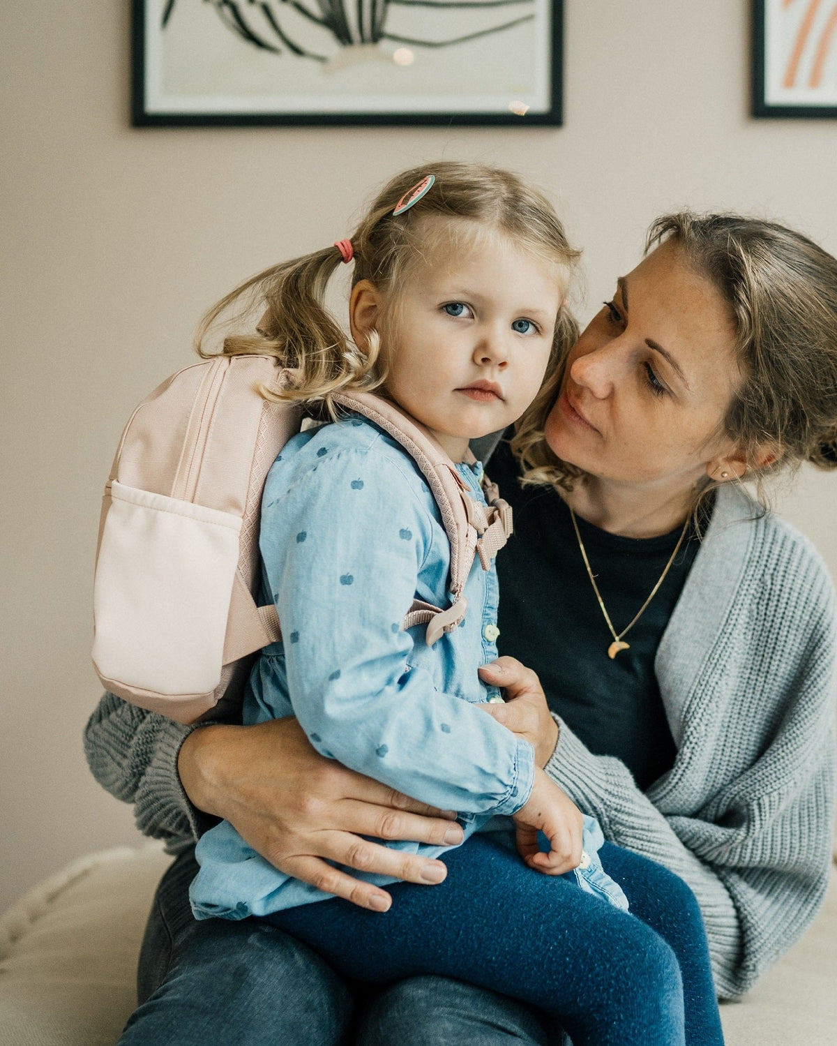 Kleines Mädchen mit rosa Kinderrucksack 'Leo' sitzt auf dem Schoß einer Frau in grauem Strickcardigan