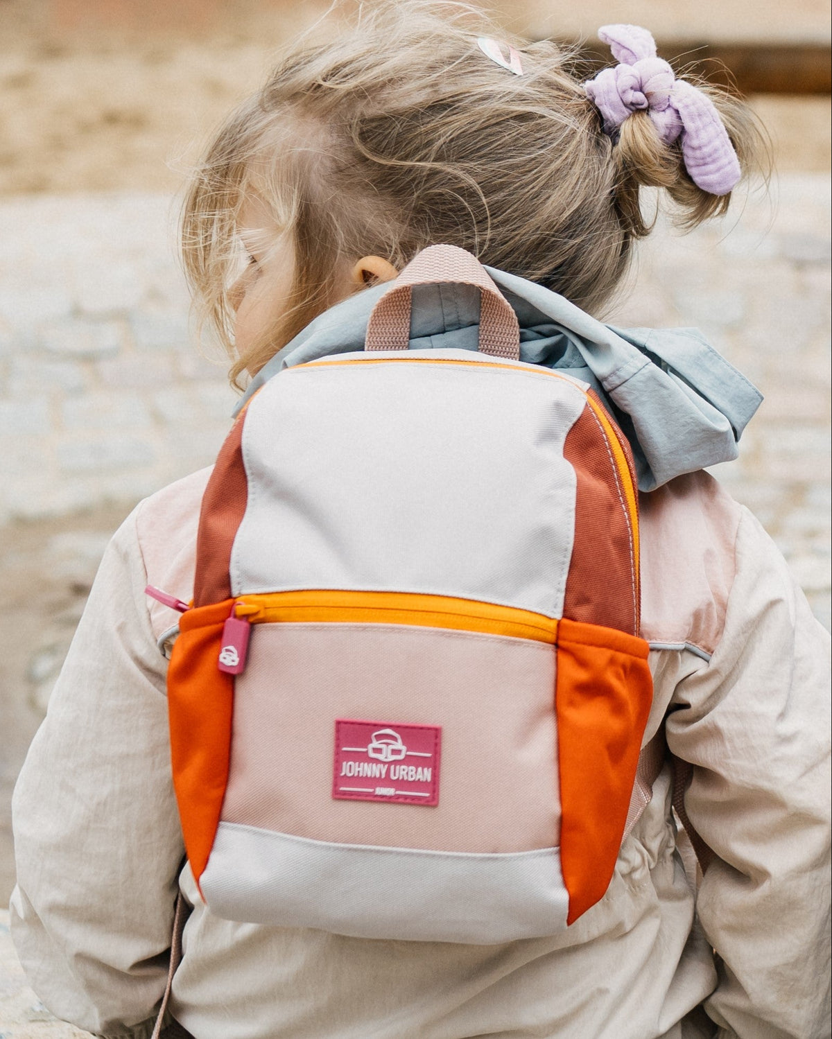 Kinderrucksack 'Leo' in Beige, Orange und Braun getragen von einem Mädchen mit hellbraunem Haar und lilafarbenem Haargummi