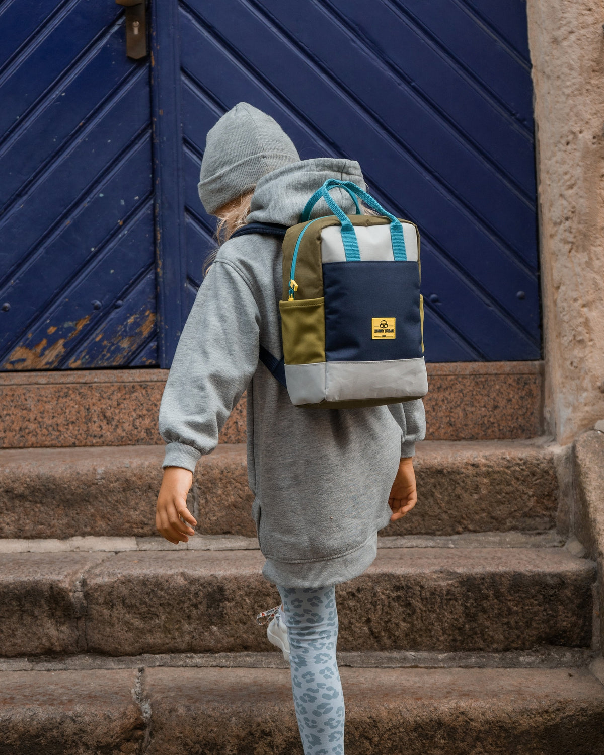 Kinderrucksack 'Linus', rechteckig in Blau, Grün und Grau, auf dem Rücken eines Kindes mit grauer Kapuzenjacke und Mütze