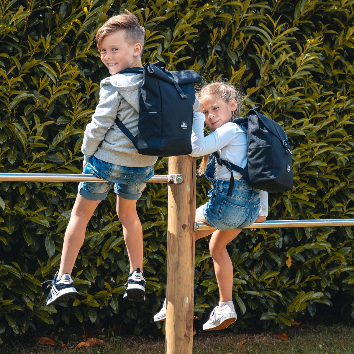 Zwei Kinder sitzen auf einem Holzgeländer, beide tragen schwarze Kinderrucksäcke 'Liam' von Johnny Urban