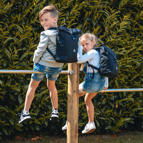Zwei Kinder sitzen auf einem Holzgeländer, beide tragen schwarze Kinderrucksäcke 'Liam' von Johnny Urban